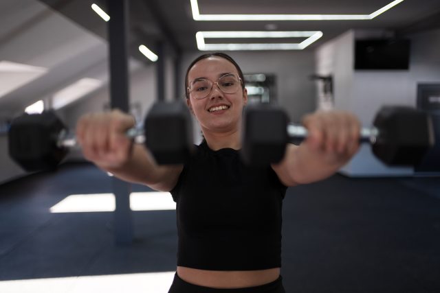 A young woman, wearing glasses and a black workout outfit, lifts dumbbells with a smile in a well-lit gym