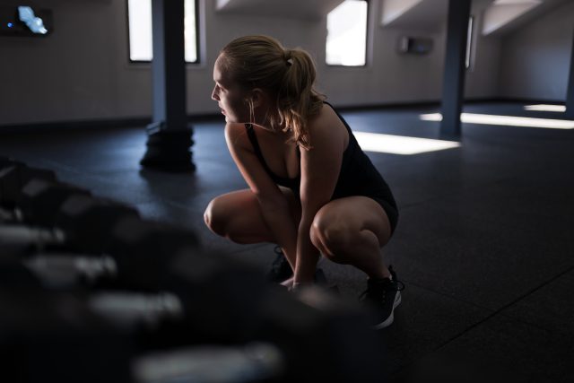 A woman in athletic wear crouches closely to a set of dumbbells in a well-lit gym