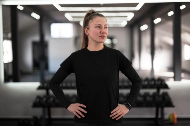 A woman stands with hands on hips, exuding confidence in a well-lit gym filled with exercise equipment