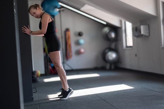 A woman stretches her calves by leaning against a wall in a bright gym
