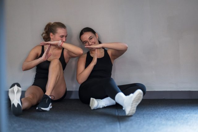 Two friends sit on a gym floor, sharing a fun moment and laughter after completing a challenging workout
