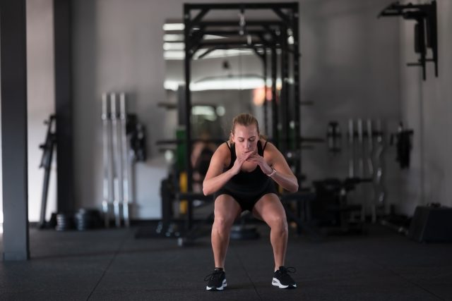 A woman exercises indoors, focusing on squats in a well-equipped gym
