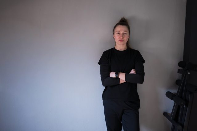 A confident individual stands with arms crossed in a stylish black outfit against a plain gray wall