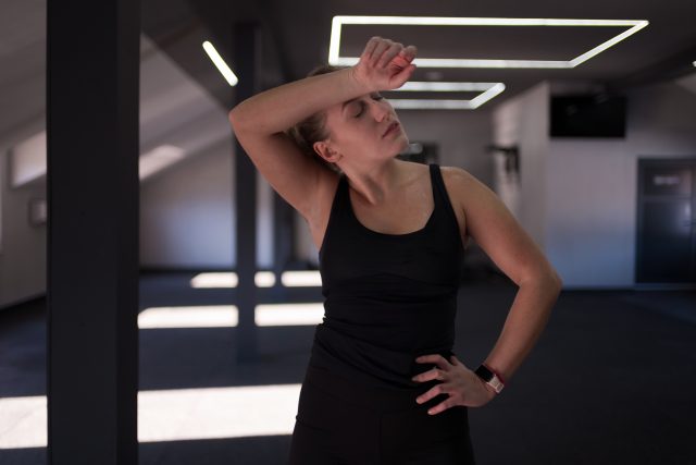 A woman wearing a black tank top stands in a fitness center, wiping her forehead after an intense workout session