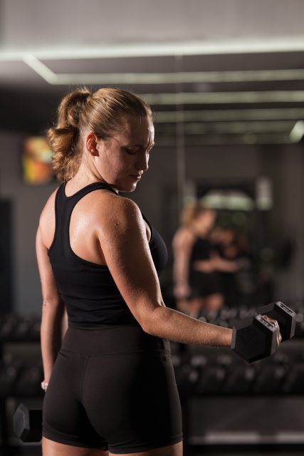 A woman in a black tank top and shorts exercises with a dumbbell in a stylish gym