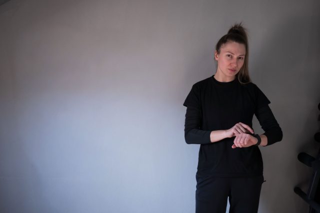 A young woman wearing a black outfit is standing near a gray wall, checking her smartwatch