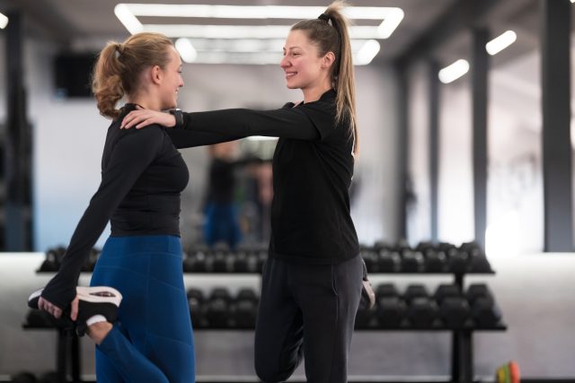 Two women stand facing each other, smiling and holding each other while stretching their legs in a bright gym