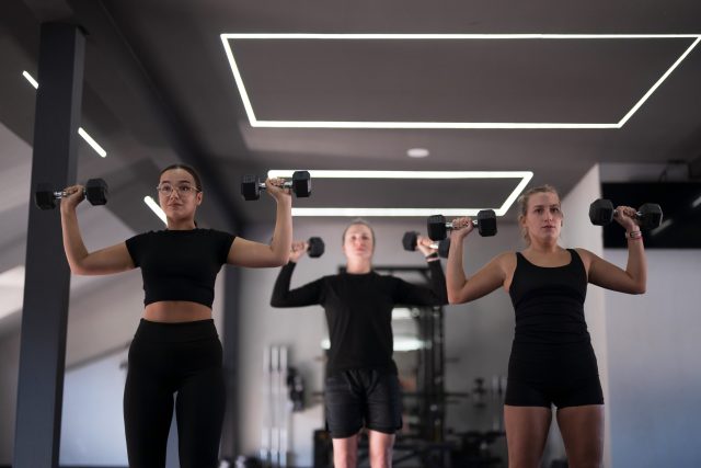 Group of women participate in a fitness class in a sleek gym, lifting dumbbells above their heads while focused on their workout