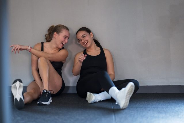 Two friends sit on the gym floor, laughing and chatting after an intense workout session