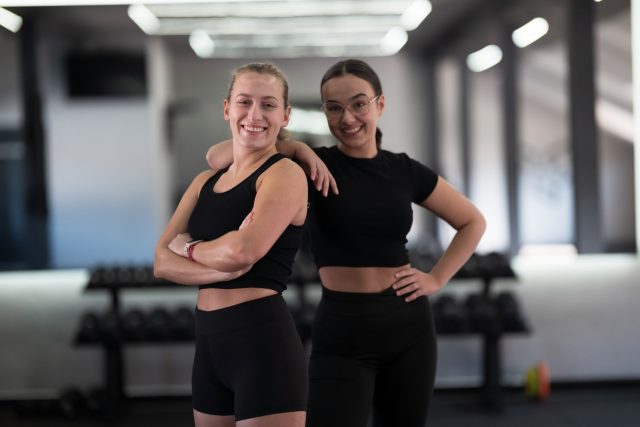 Two women stand next to each other in a gym, smiling and posing in matching black workout clothes