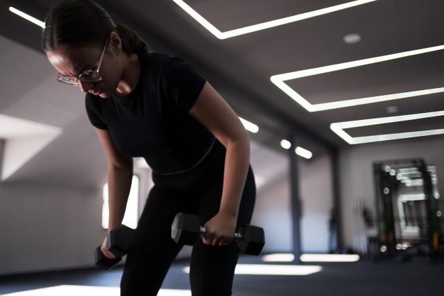 A young woman performs a strength training routine with dumbbells in a well-lit gym