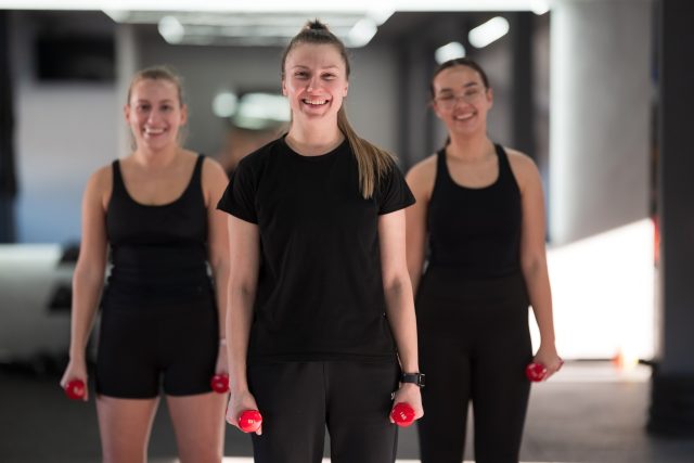 In a well-lit gym, three women smile while holding red dumbbells, showcasing their enthusiasm for fitness