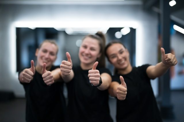 Group of women wearing matching black shirts show thumbs up with smiles in a bright gym