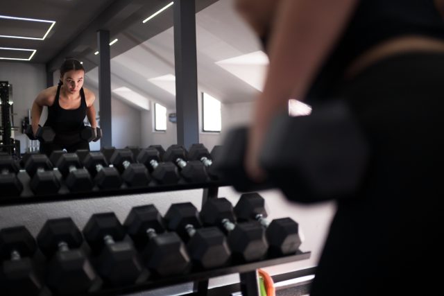 A woman is engaged in weight lifting at a gym, concentrating on her form as she looks into the mirror