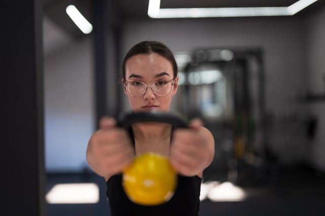 A dedicated young woman lifts a yellow kettlebell in a stylish gym, demonstrating focus and determination as she works on her strength…