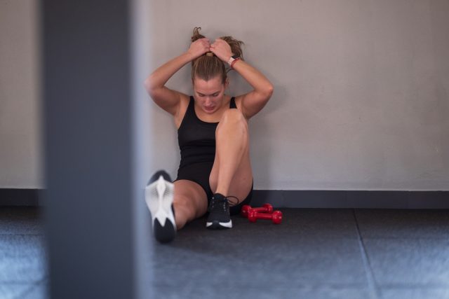 A woman sits against the wall in a gym, resting after an intense workout