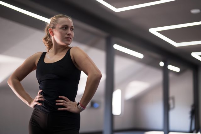 In a spacious fitness studio, a female athlete stands poised with her hands on her hips, showcasing a determined expression while preparing…