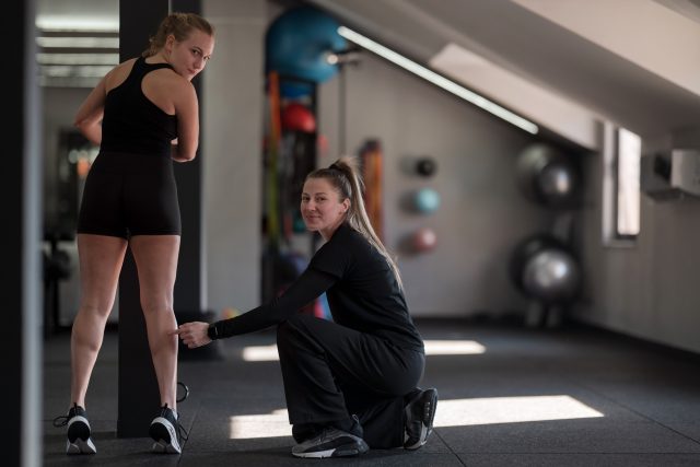 In a modern gym, one woman kneels to adjust the shorts of her workout partner