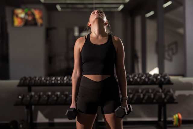 A woman stands in a well-lit gym lifting dumbbells in both hands