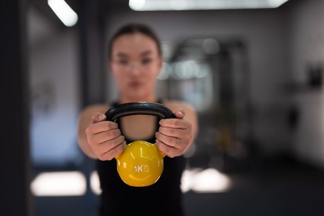 A woman holds a yellow kettlebell in a gym, focusing on her workout routine in the late afternoon