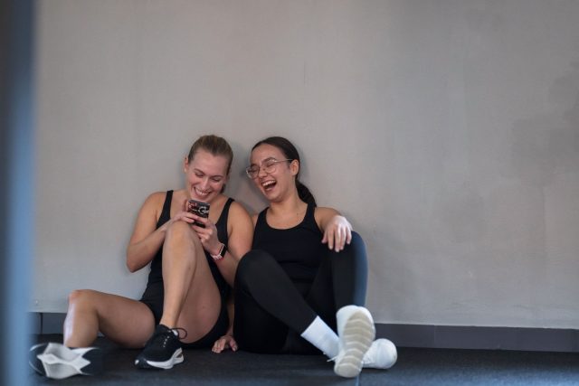 Two women sit on the gym floor, taking a break from their workout