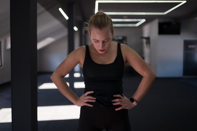 A woman in a black tank top stands with her hands on her hips, catching her breath after a hard workout in a spacious fitness studio