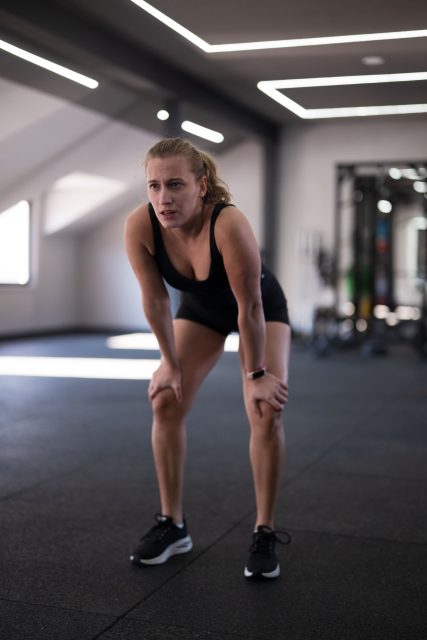 A woman in workout clothes takes a break in a stylish gym, showing signs of fatigue after an intense training session