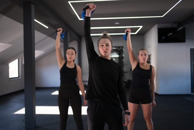Group of women participate in a group workout, lifting weights in a spacious gym with modern design elements and ample sunlight