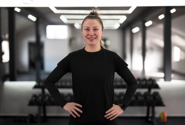 A woman dressed in a black workout outfit smiles while standing with hands on hips in a sleek gym