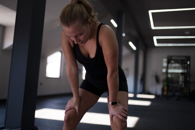 A woman in a black workout outfit leans forward, resting her hands on her knees