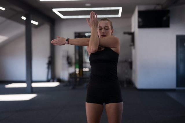 A young woman performs a stretching routine focusing on her upper body in a contemporary gym
