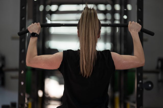 In a well lit gym, a woman with a ponytail performs a pull-up on a bar