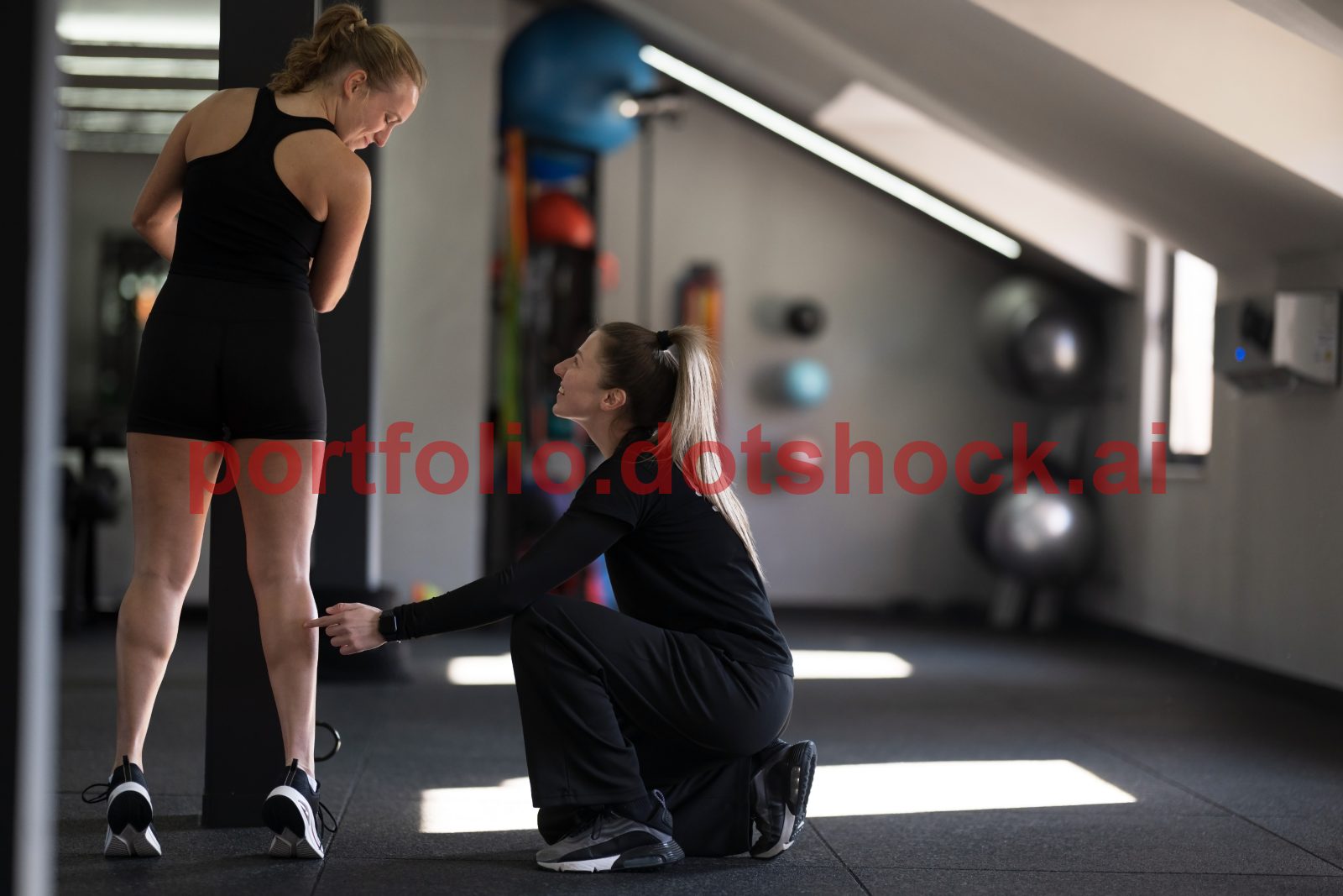 A fitness trainer is crouching beside a woman in workout gear, correcting her leg exercise technique in a well-lit gym
