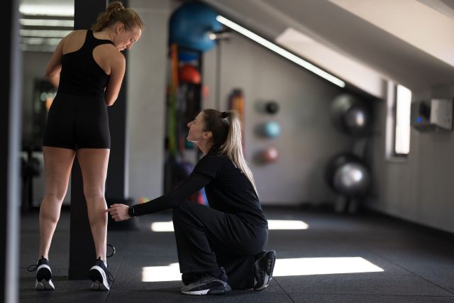 A fitness trainer is crouching beside a woman in workout gear, correcting her leg exercise technique in a well-lit gym