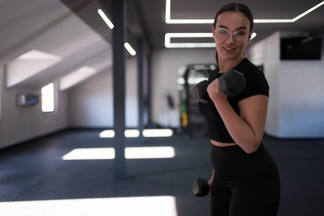 A fit young woman exercises with dumbbells in a bright, spacious gym