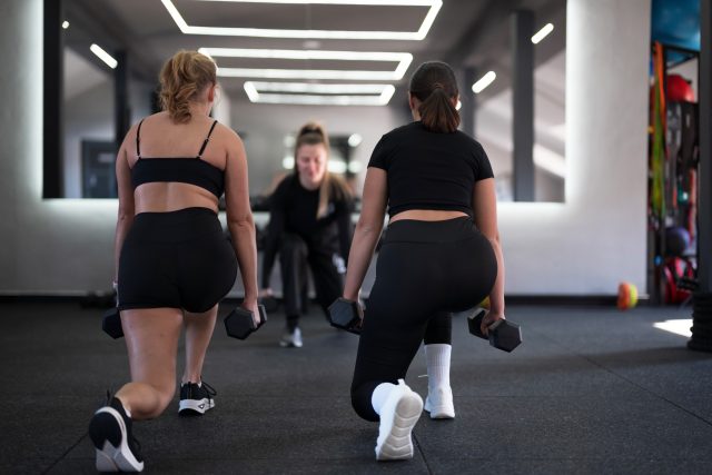 Two women are engaged in lunges while holding dumbbells in a contemporary gym setting