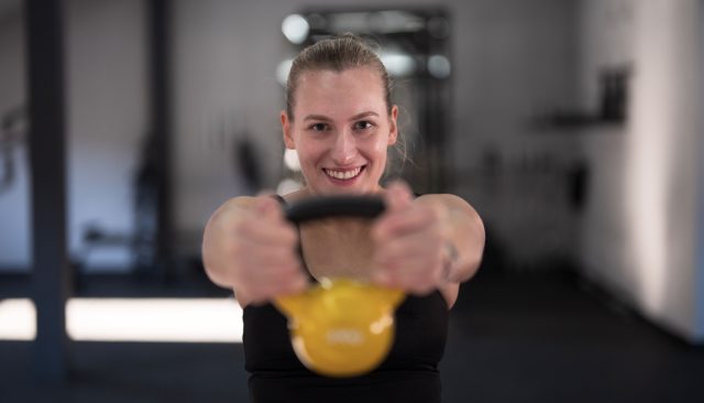 A woman holds a yellow kettlebell in front of her, smiling confidently in a gym