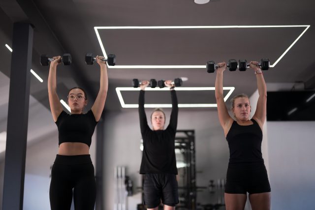 Group of women are exercising in a stylish gym, each lifting dumbbells above their heads