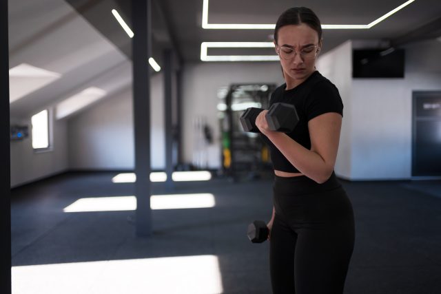 A woman focuses on lifting a dumbbell in a bright, spacious gym