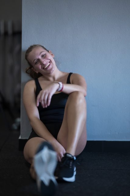 A young woman sits on the floor against a wall, smiling brightly after completing her workout