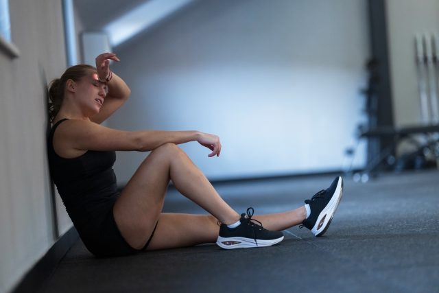 A woman sits against a gym wall, looking tired after finishing her workout