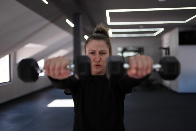 A determined woman works on strength training by lifting dumbbells in a spacious gym with bright, natural light coming through the windows